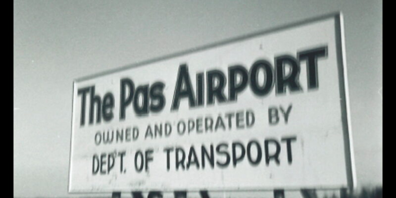 A man stands under a sign that reads, "The Pas Airport Owned and Operated by Dept. of Transport"