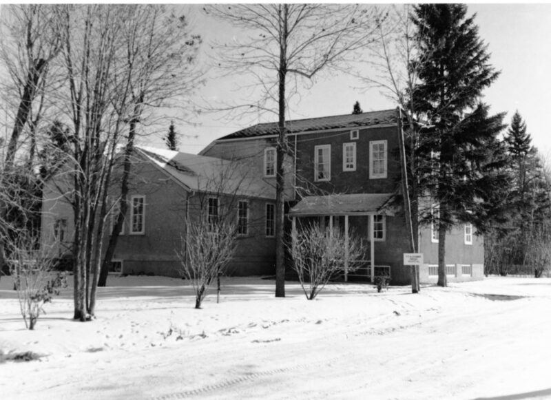 A black and white photo of an older but large two-story building. In the background is a smaller, white two-story house. The buildings are surrounded by trees.
