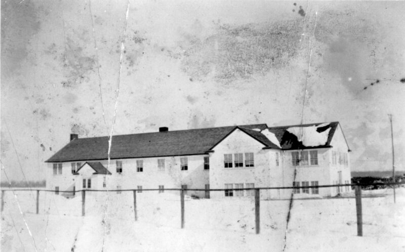 Black and white photo of a long, rectangular two-story white building. There is snow on the ground. A fence made of wood and barbed wire crosses in front of the building in the foreground of the photo.
