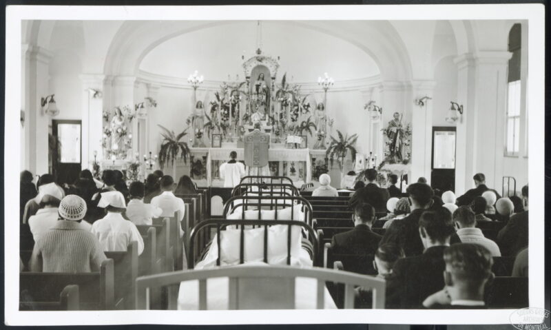 A view from the back of a chapel. People sit in the pews facing the large altar at the front. A row of hospital beds lines the aisle between the pews.