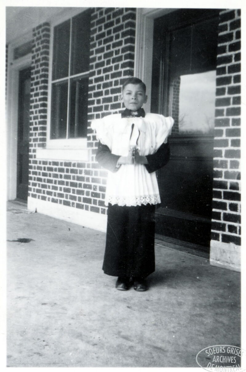 A boy in a white lace top stands outside of a building with his hands in a prayer position.