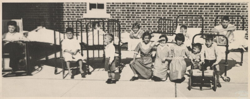 A group of children outside near a brick building. Some children are in hospital beds, some are in wheelchairs.