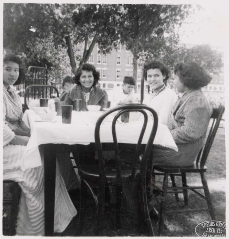 Four young women sit at a table outdoors. A boy can be seen at another table behind them.