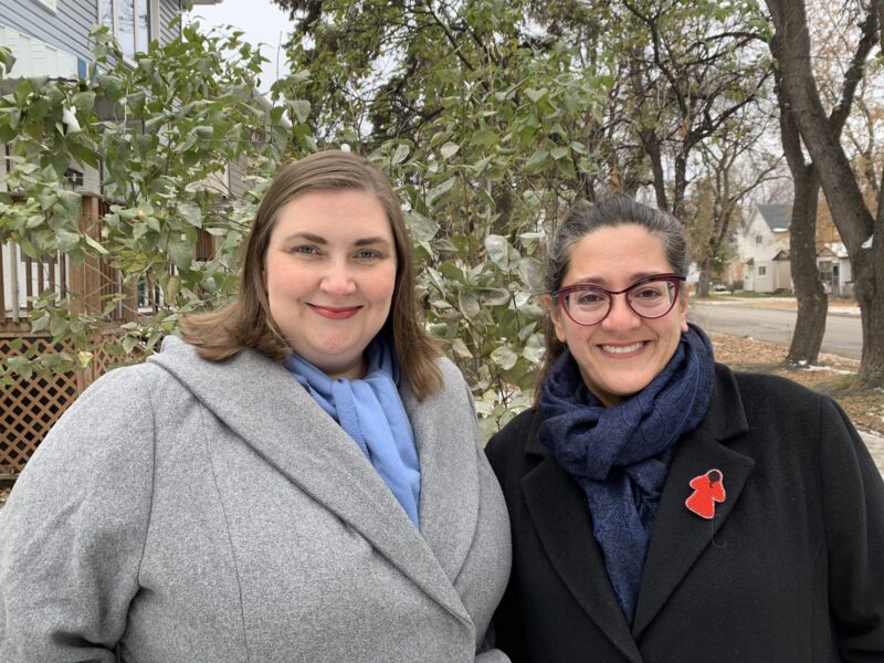 Two women standing next to each other in front of a tree. The woman on the left wears a grey coat and the woman on the right wears a black coat.