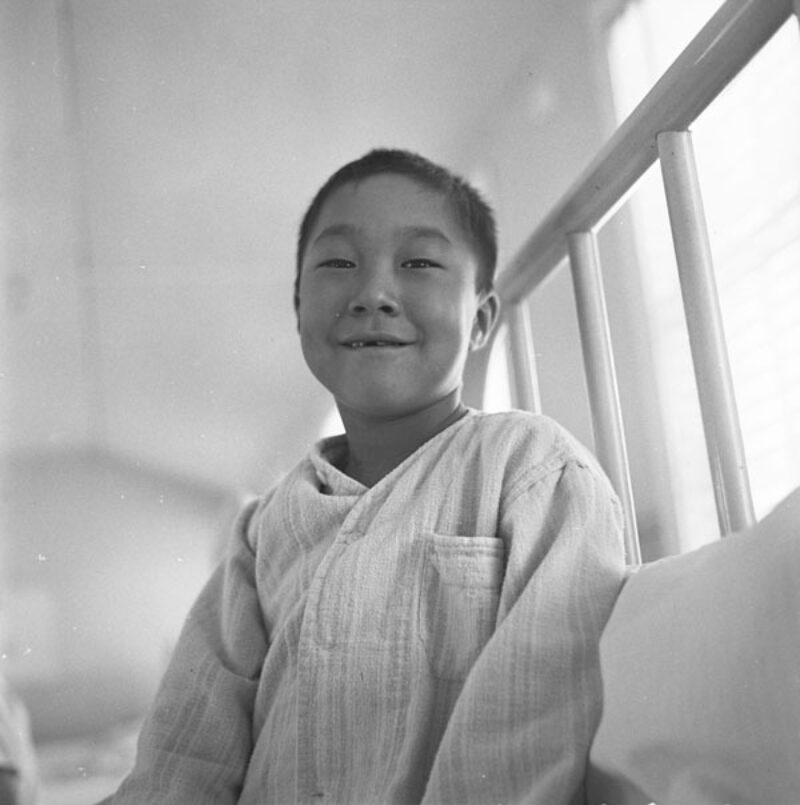 An Inuk boy sits on a bed and smiles.