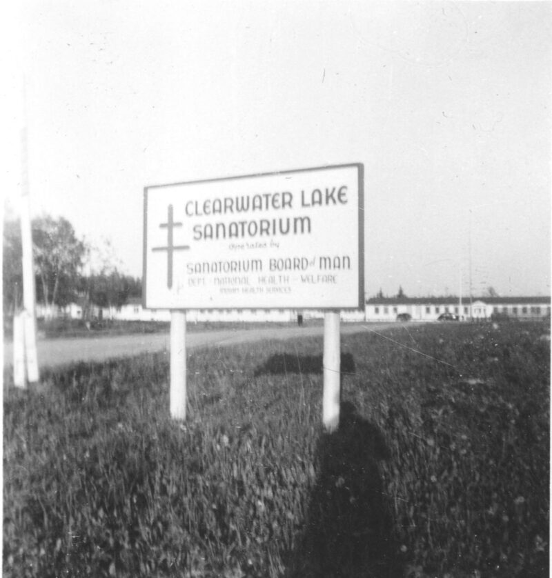 A sign next to a road reads: "Clearwater Lake Sanatorium // Sanatorium Board of Man." The road leads to a long single-storey building.
