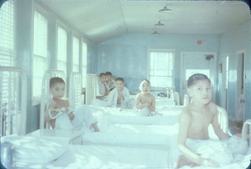 Six boys sit in hospital beds. Their hair is buzzed short, and most of the boys are not wearing shirts.