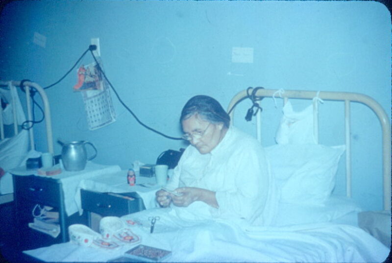 A woman does beadwork in a hospital bed.