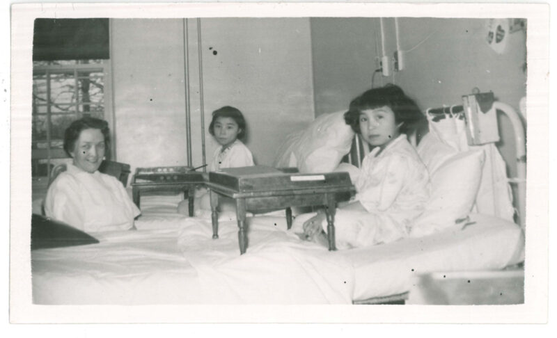 Two girl patients in beds with lap writing desks, with a nurse sitting between them