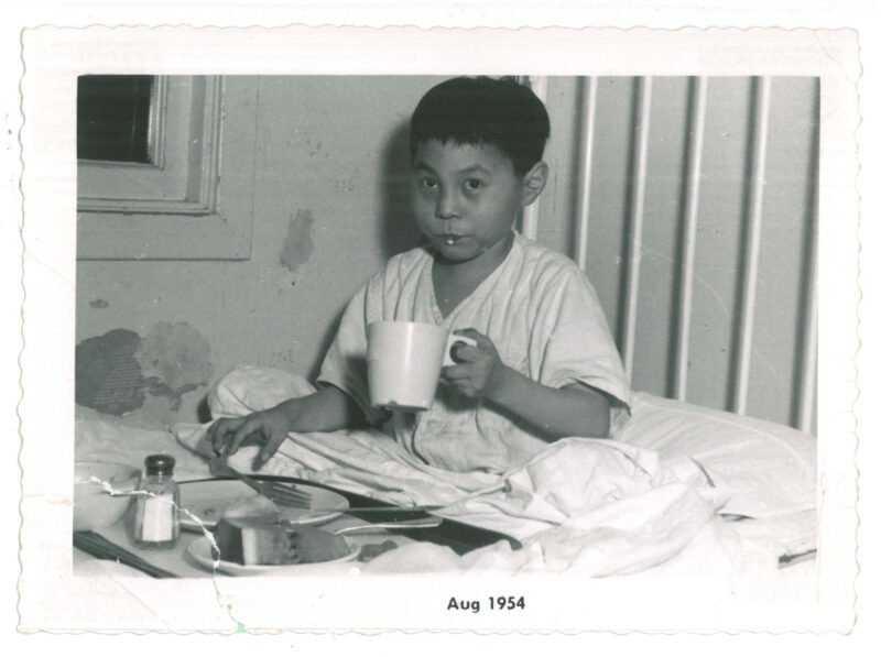 Boy patient in bed holding a mug, with a food tray on the bed in front of him. The wall is peeling next to him.