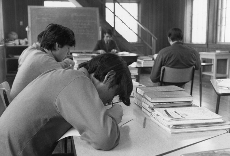 Six young men writing at desks. The young man closest to the camera is hunched over, writing on a paper. Stacks of books sit along the desks, and a chalkboard stands in the background.