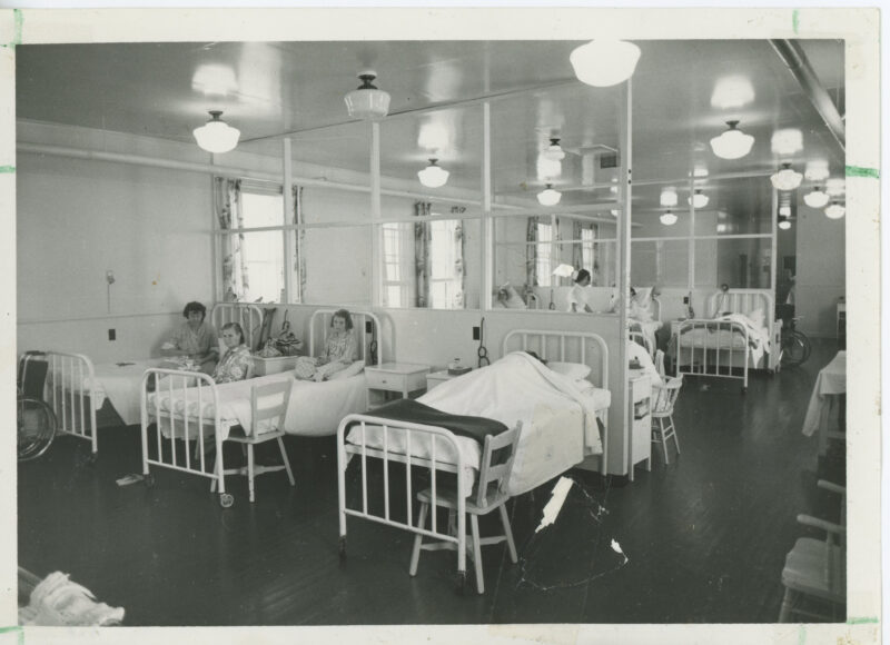 A view of a hospital ward. The ward partitioned, and beds are lined up against the partitions. Female patients can be seen sitting in beds throughout the ward, and a nurse stands in the background.