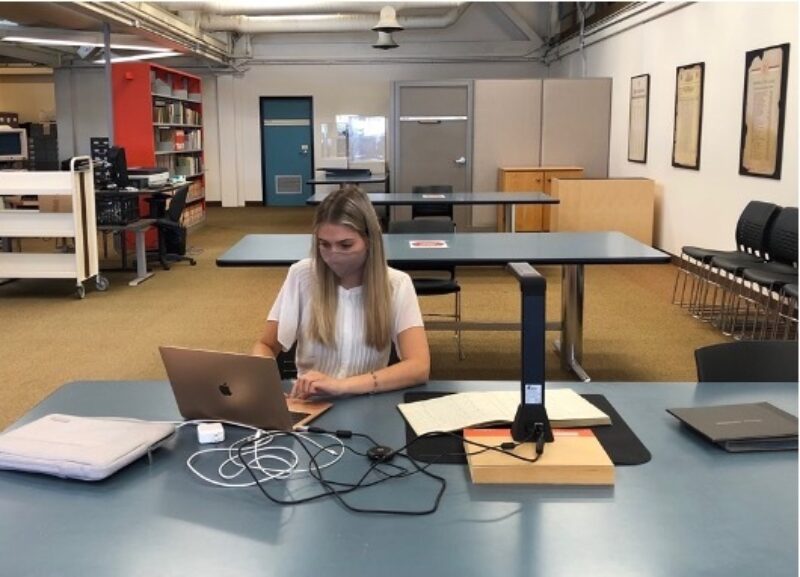 A young woman with long blond hair, wearing a mask, sits at a table. On the table are some documents and a scanning device. The woman is working on a laptop. There are bookshelves in the background.