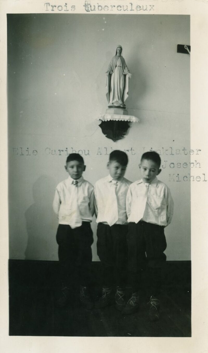 Three young Indigenous boys wearing matching dark pants and white shirts, lined up in a row.