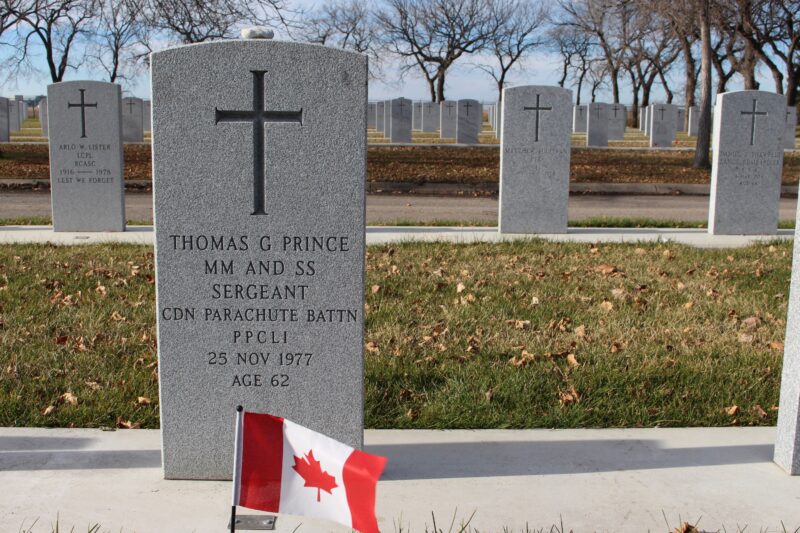 Photo of Sergeant Tommy Prince's military headstone with a small Canadian flag placed in front. The headstone is grey with a cross engraved at the top. The headstone text reads "Thomas G Prince, MM and SS, Sergeant, CND Parachute Battn, PPCLI, 25 Nov 1977, Age 62."