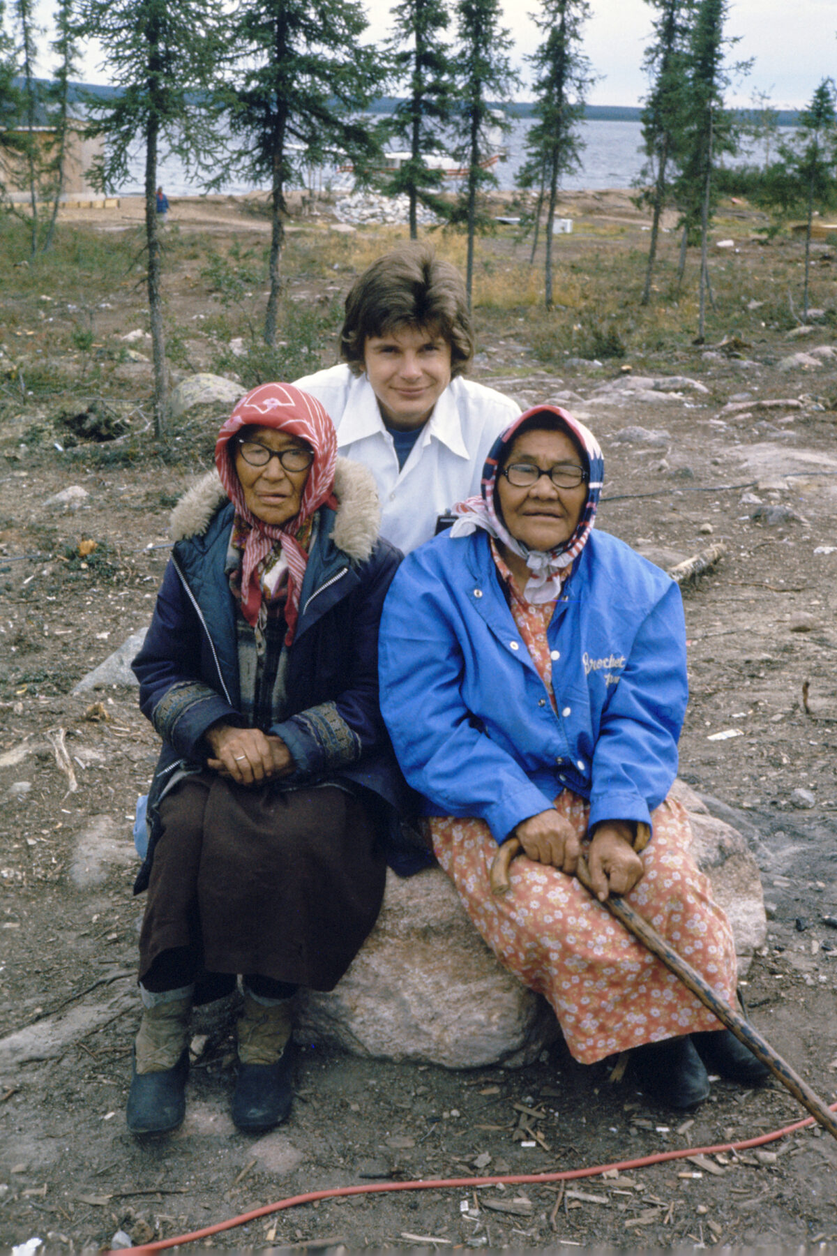 Two elderly women sitting on a rock outside by a lake, with another person crouched behind them.