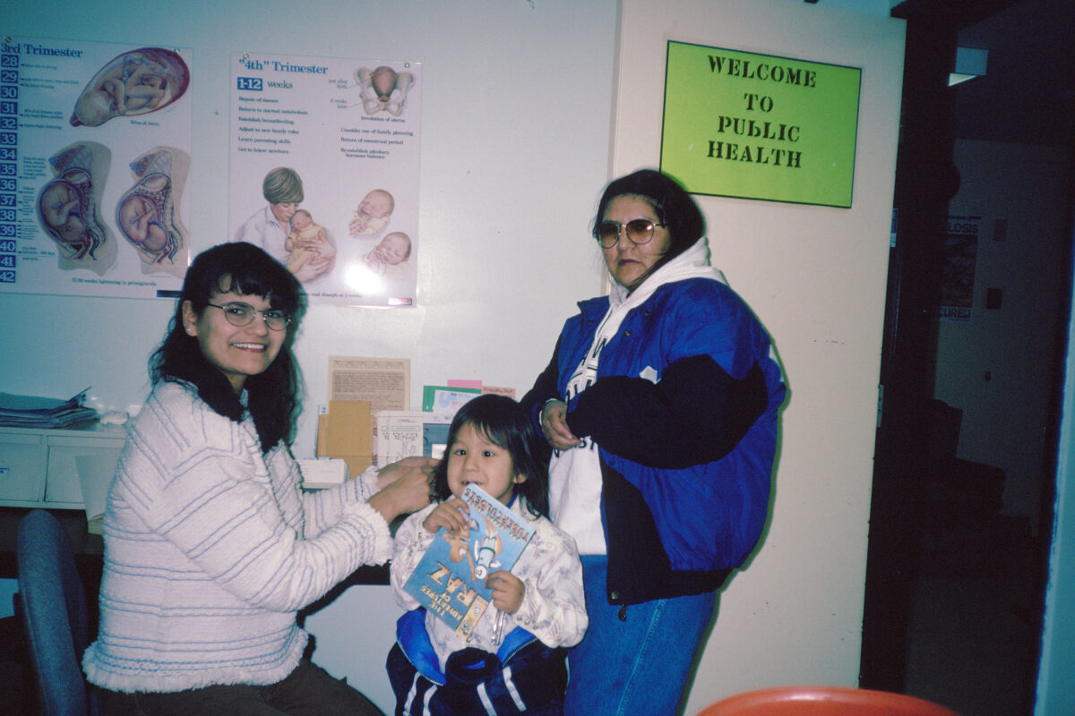 Two women and a child in a doctor's office. The child chews on a magazine called "The Adventures of Raz: Raz Learns About Tuberculosis." A sign on the wall says "Welcome to Public Health."