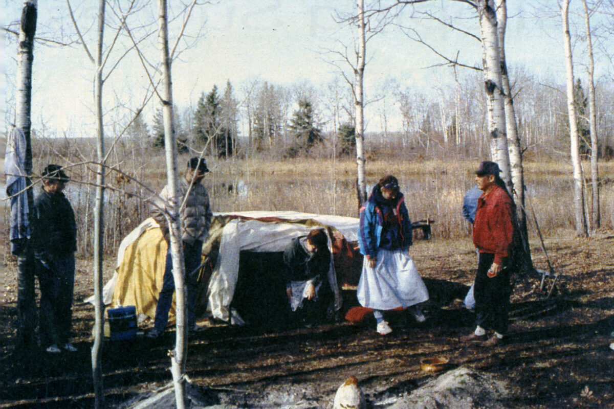 Four people standing next to a sweat lodge, and one woman exiting the sweat lodge.