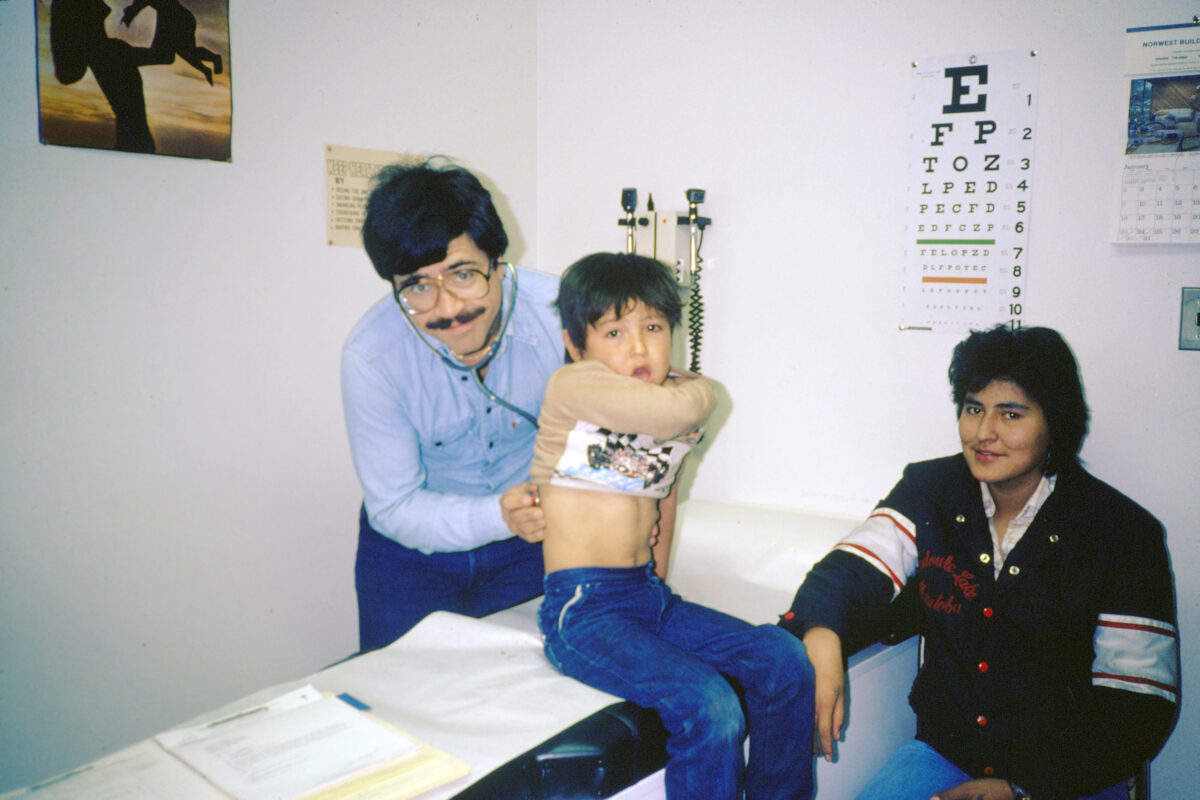 A boy lifting his shirt while a man listens to his back with a stethoscope. Another person sits next to the boy.