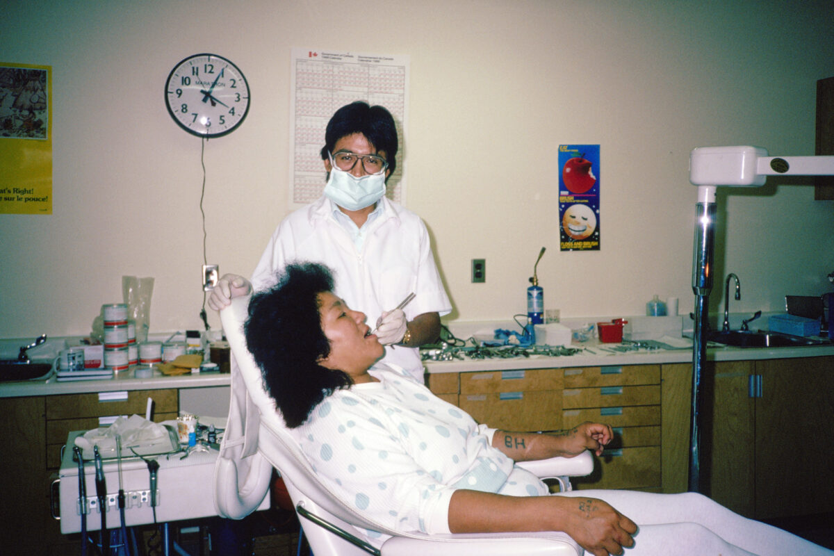A woman in a dentist's chair, with the dentist standing beside her holding an implement in her mouth.