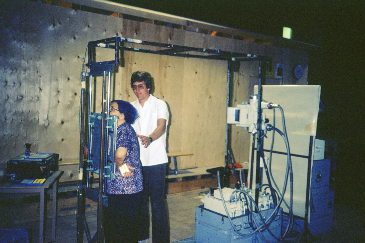 A man guiding a woman as she presses her chest against an x-ray plate.