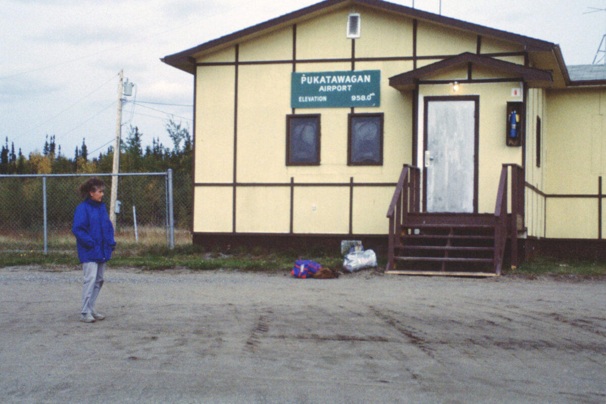 A woman in a blue coat, standing next to a building with a sign that reads, "Pukatawagan Airport // Elevation 958.0"
