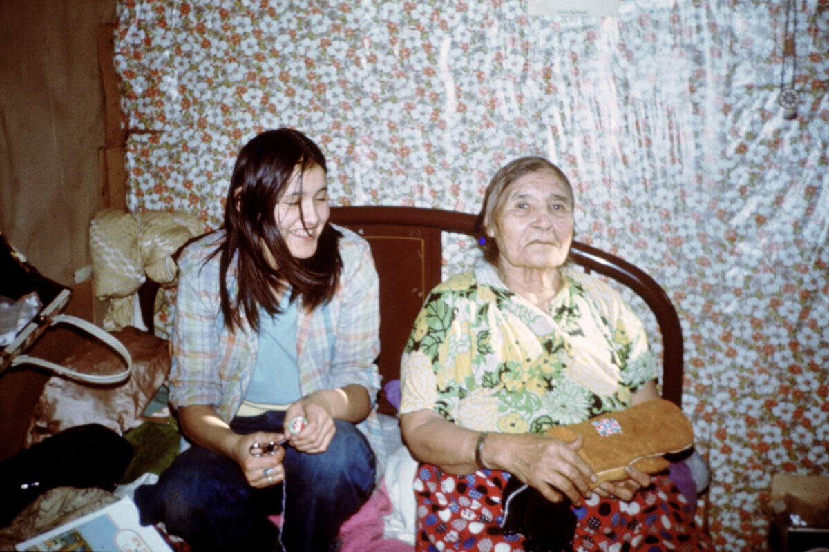 A young woman/teen sits lets to an elderly woman holding a beaded leather mitten.
