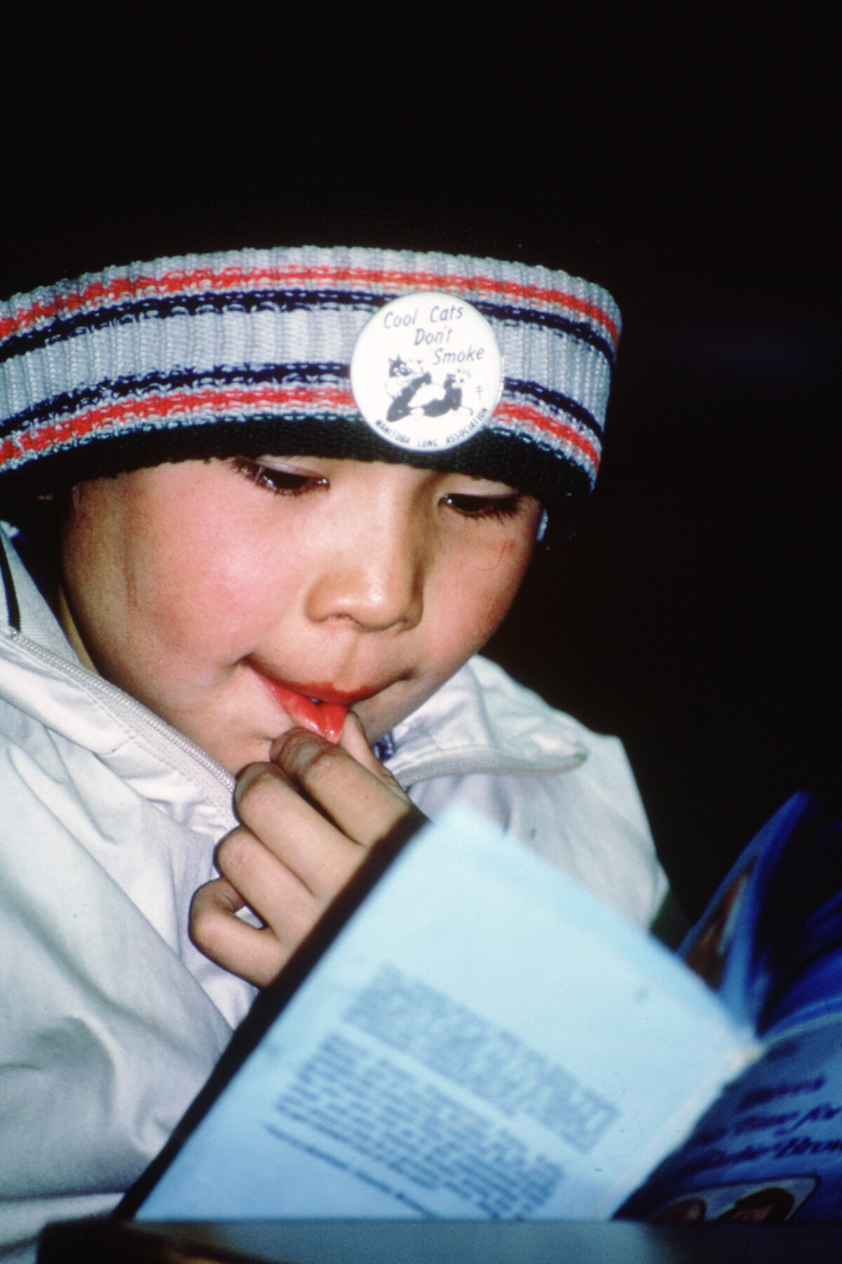 A young child holds their bottom lip while reading a book. A sticker on their hat reads, "Cool Cats Don't Smoke"
