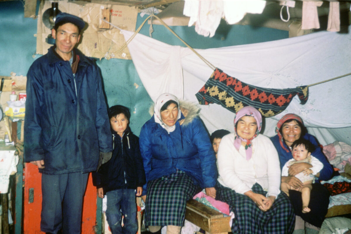 A man and a boy stand beside three women and two other children sitting. Blankets hang from ropes above them.