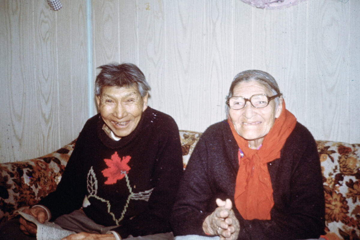 An elderly couple sit on a flowered couch in a wood-panelled room. They both smile at the camera.