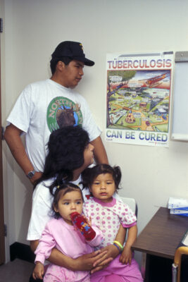 A family in a doctor's office, looking at a poster on the wall that reads "Tuberculosis Can be Cured." Two young children sit on a woman's lap, and a man stands behind them.