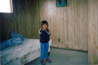 A smiling boy holds something next to his face. He stands in a wood-panelled room next to a bed.