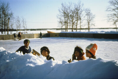 Four children peering over a snow wall bordering a skating rink.