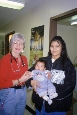 A woman with a stethoscope around her next standing next to a woman holding an infant