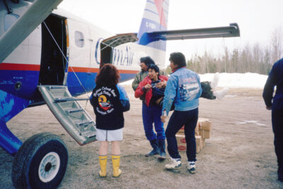 Two men carrying another man onto a "Calm Air" airplane, while a woman watches.