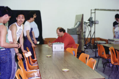 Four young men standing in a line along a table with orange chairs. A man sits at the end of the table, writing on a paper.
