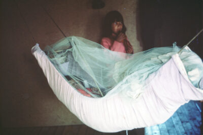 A child in a hammock covered in mesh, and another child stands beside, holding something to her mouth.