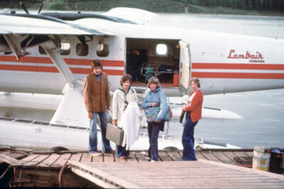 Four people next to a Lambair seaplane holding luggage.