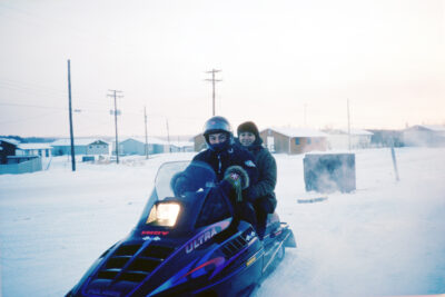 Two people on a snowmobile. Small buildings stand in the background.