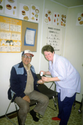 A nurse bends over to hand medicines and a small cup to a seated man. They both smile at the camera.