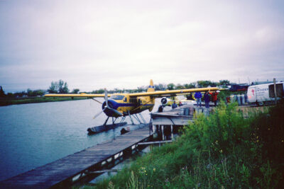 A yellow seaplane sits at a dock. A group of people stand on the dock next to the plane.