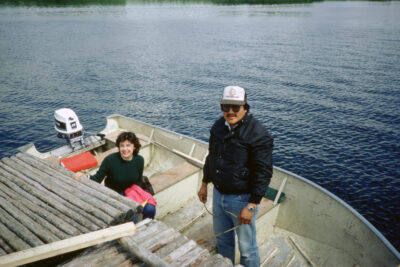 A man stands in a boat parked at a dock, and a woman sits in the boat with one hand on the dock.