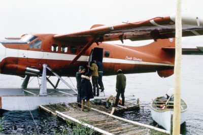A group of people boarding a seaplane. A white canoe sits on the water next to the plane.
