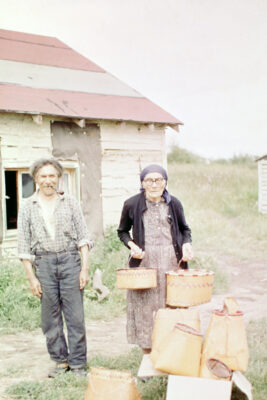 A man with a pipe in his mouth next to a woman holding two birch bark baskets. They stand outside next to a house.