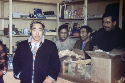 A man looks at the camera in a co-op store, while three men stand on the other side of the counter and rummage through boxes.