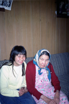A teenager sits on a couch next to an elderly woman in a wood-panelled room.