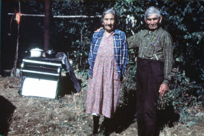 An elderly couple stands next to an outdoor stove. The man has his arm around the woman's shoulder.