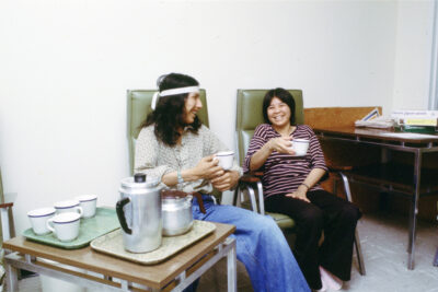 Two people sitting on green chairs laughing, holding mugs. A tray of mugs and coffee carafes sits next to them.
