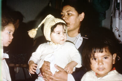 A baby sits on a woman's lap, and two young children stand next to her.