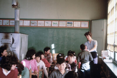 A boy stands at the back of a classroom with a woman. She appears to be fixing his collar or pinning something to his shirt. Other students sit at their desks, and some turn to watch the boy.